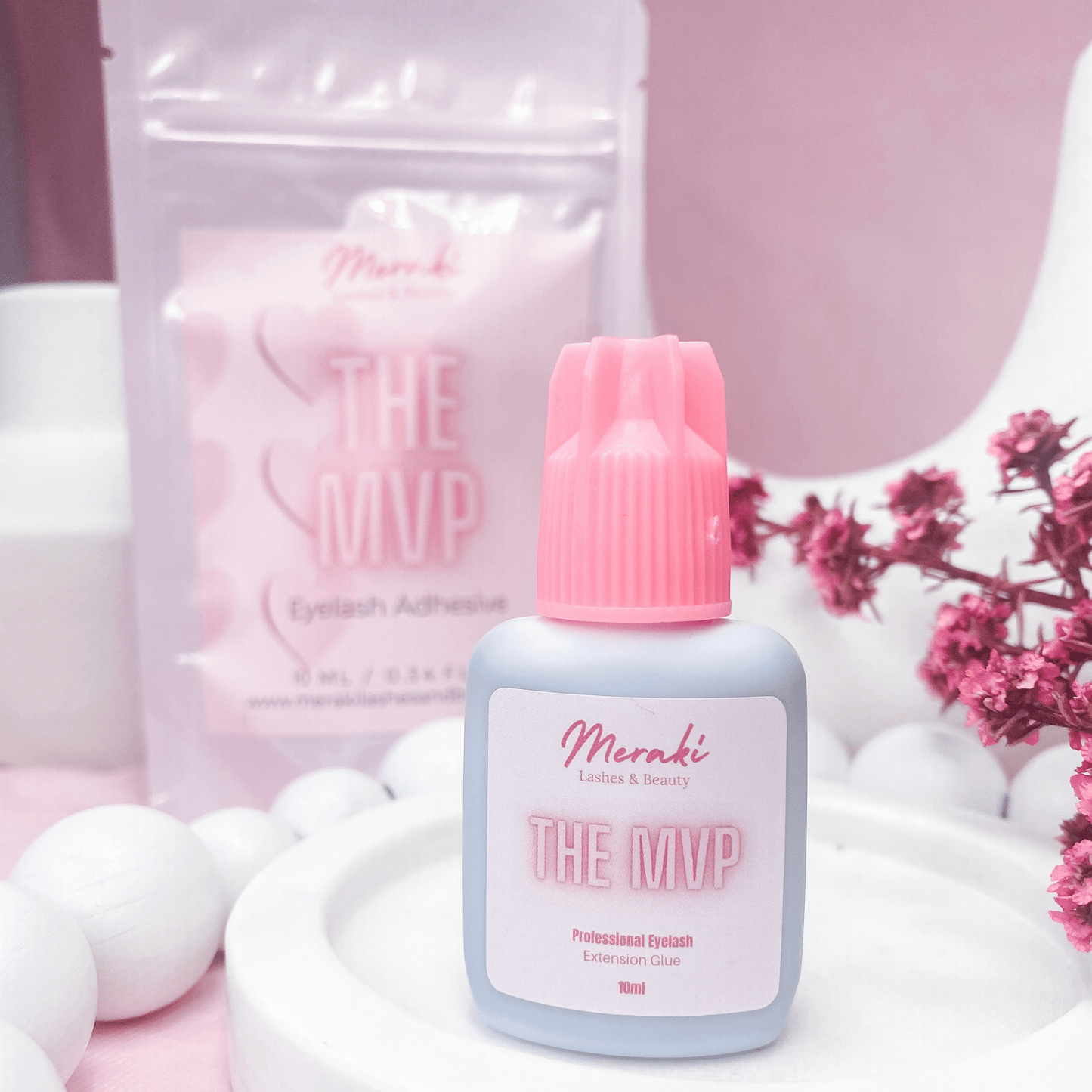 Meraki MVP lash glue bottle and packaging styled with soft pink props; clean product flat lay.